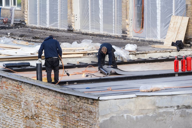Two construction workers install a black waterproofing membrane on a flat roof