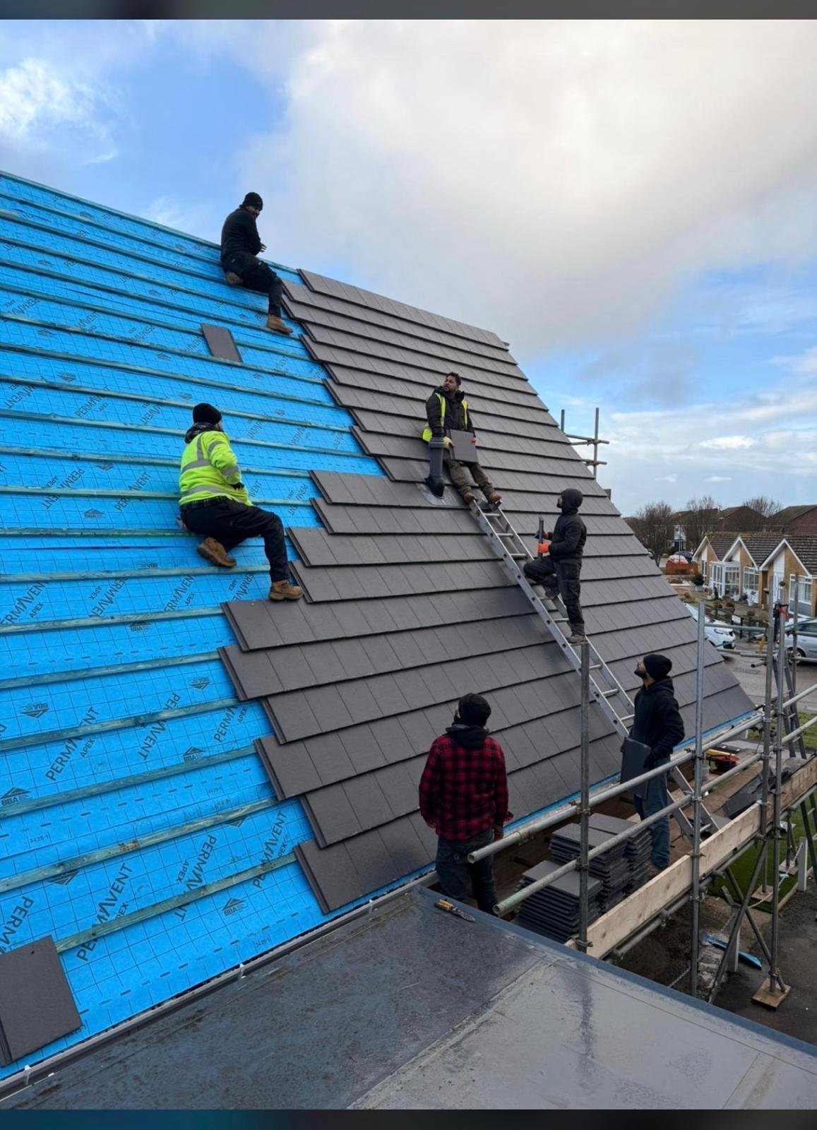 Multiple workers installing dark roof tiles on a residential home under construction, with one worker wearing a yellow vest on blue sheeting and metal scaffolding visible