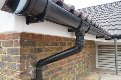 Black downspout and gutter system installed on brick house exterior below tiled roof