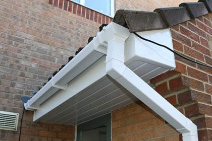 White rain gutter and downspout installed on brick building exterior with metal flashing at roof corner