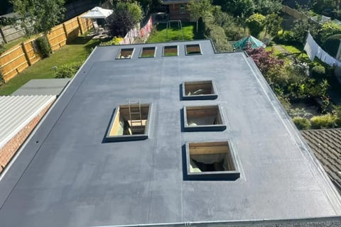 Aerial view of a flat gray roof with three square skylights and surrounding residential properties and gardens