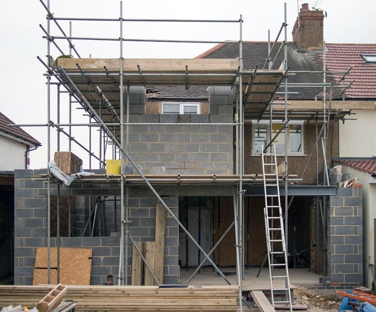 Construction site showing two-story brick house under renovation with metal scaffolding, ladders, and building materials